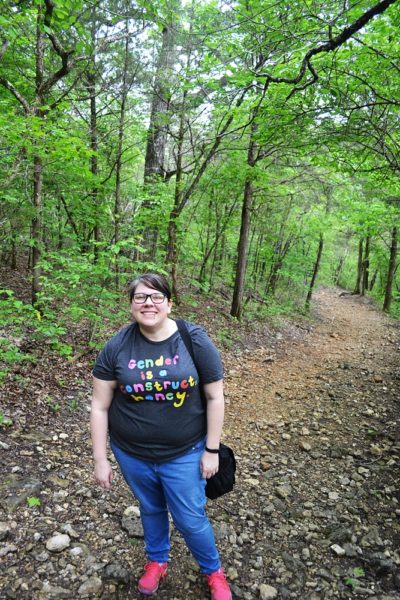 Fat woman with glasses standing on a trail, shirt reads gender is not a construct honey