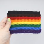 Crochet inclusive rainbow pride flag is held by a hang on a white background