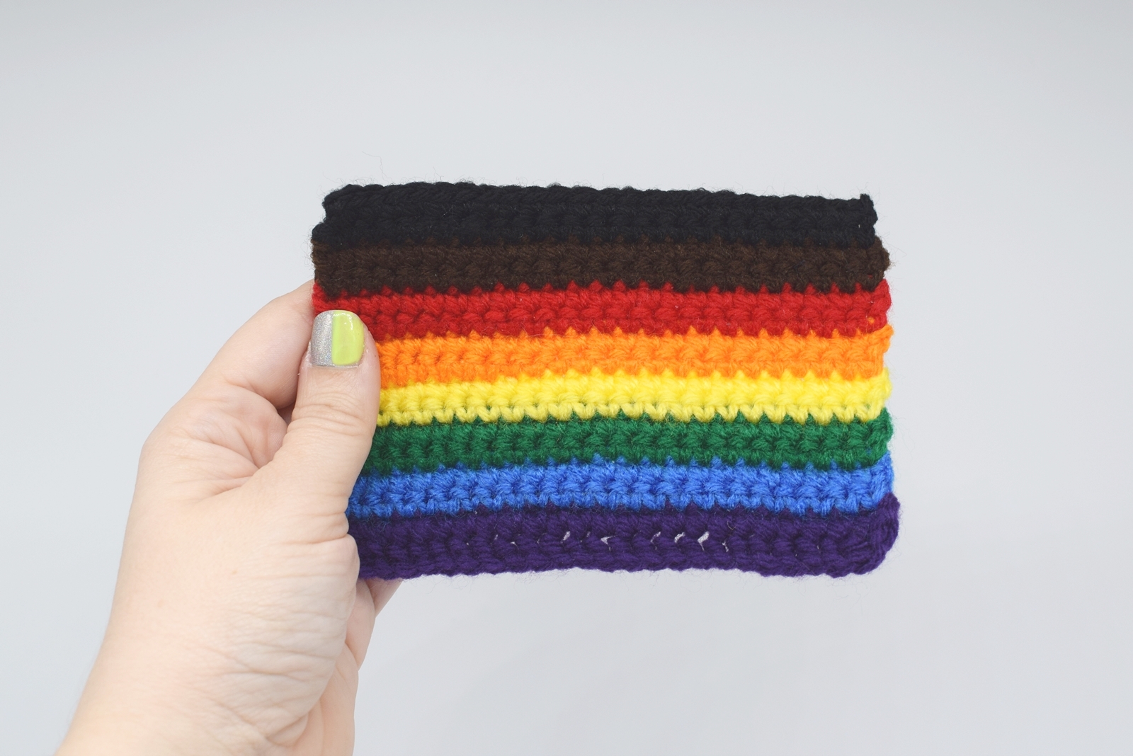 Crochet inclusive rainbow pride flag is held by a hang on a white background
