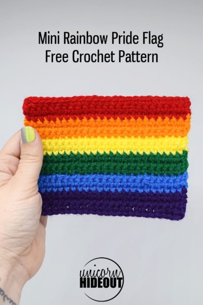 hand holds a mini rainbow Pride flag against a white background, text on the image says "Mini Rainbow Pride Flag Free Crochet Pattern, Unicorn Hideout"