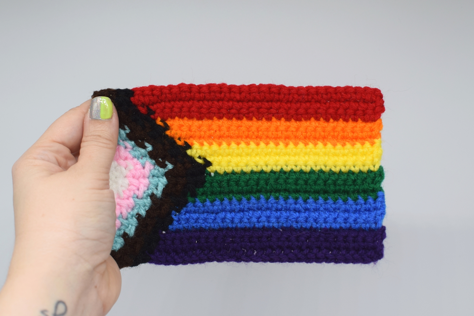 Crocheted rainbow Pride Progress flag held by a hand on a white background
