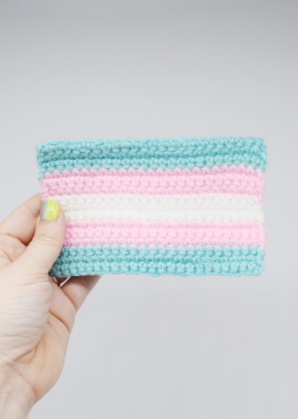 Hand holds a small crochet transgender Pride flag against white background