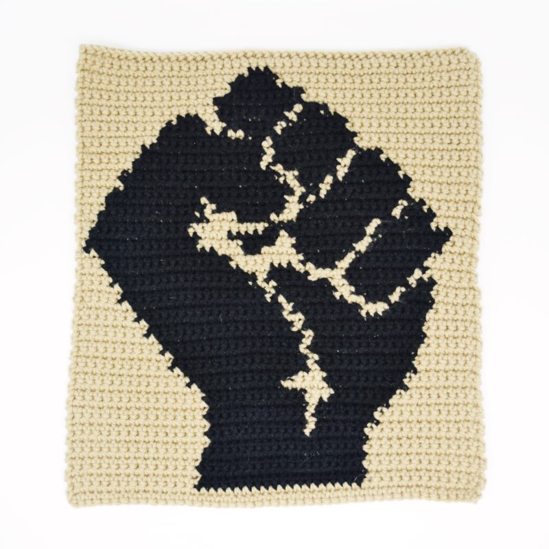 Crochet wall hanging with a black fist on a brown background.