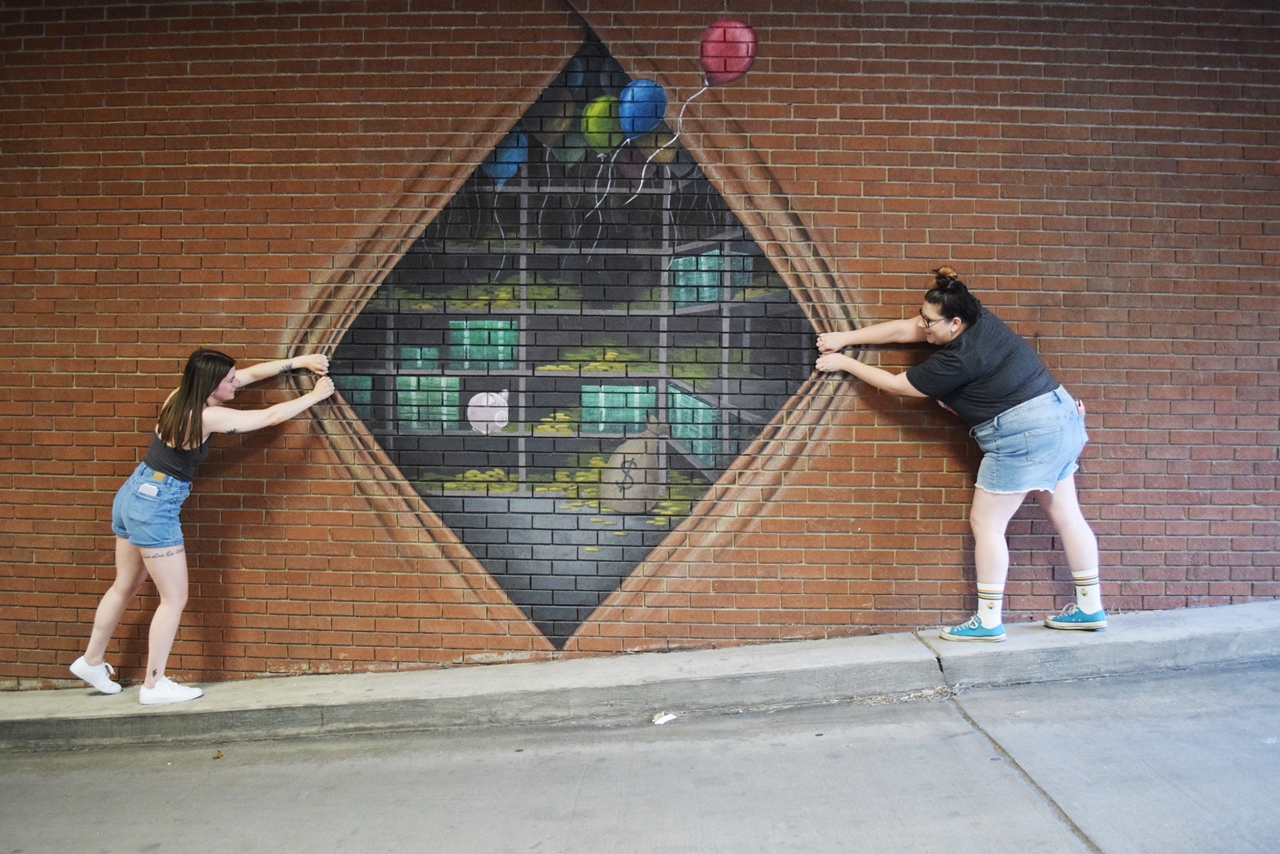 Two women stand on either side of a mural painted to look like a bank vault being pulled open