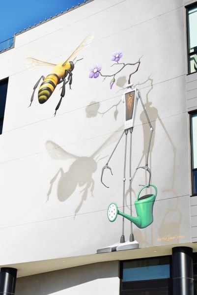 Multi story mural on the side of a gray building, a bee approaches a skinny robot with flowers for a head, holding a watering can