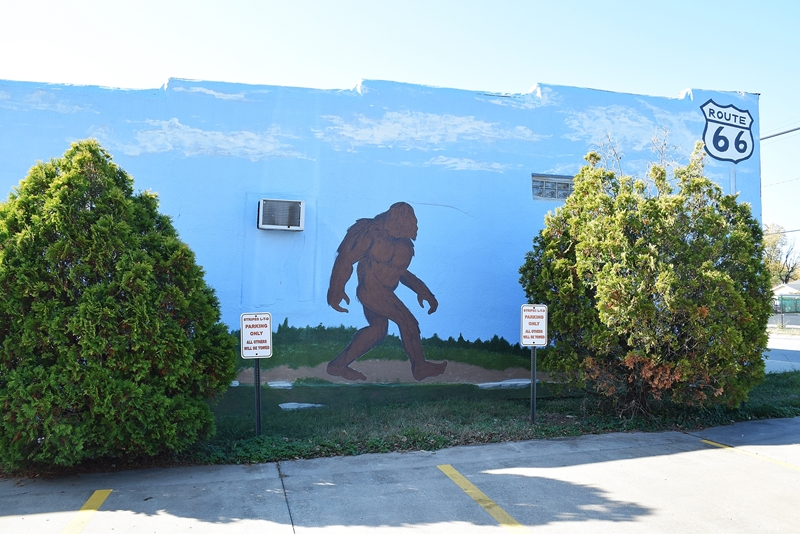 Two real bushes stand in front of a mural of a bigfoot in front of a skyline