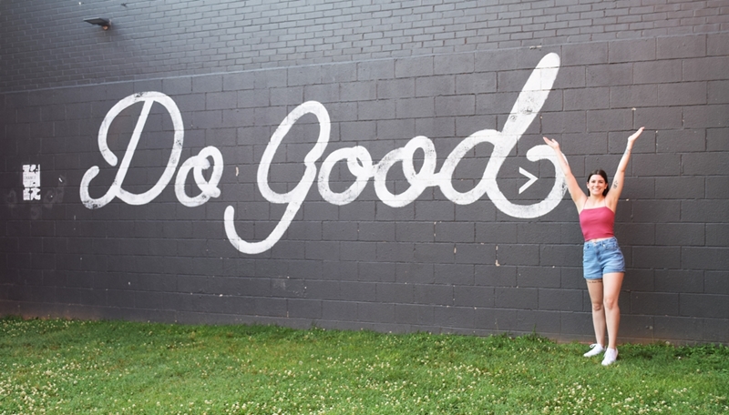 gray brick wall with Do Good painted on it, woman stands to the side with her hands up