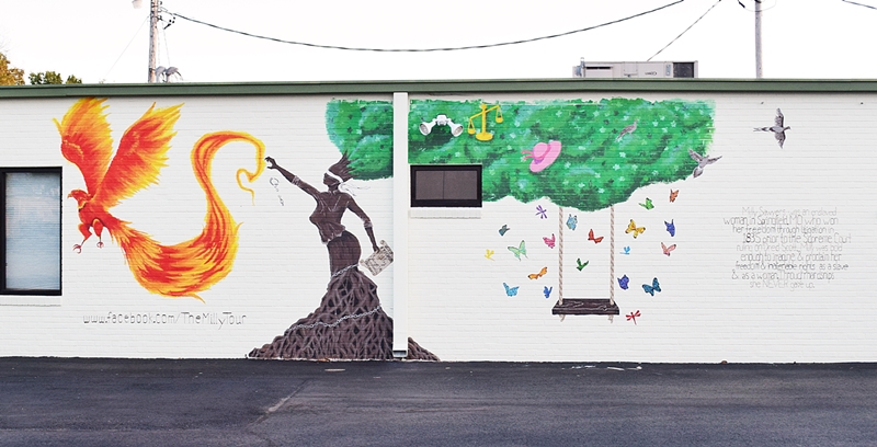 Mural on the side of a white buidling featuring a tree in the form of a woman, reaching out toward a phoenix, her hair is the leaves on the tree and a swing hangs from the tree, surrounded by butterflies