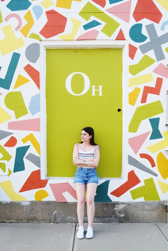 Woman stands in front of a green door, mural of shapes around the door