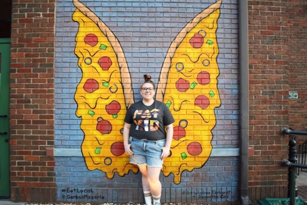 Carly stands in front of a mural of wings that look like pizza