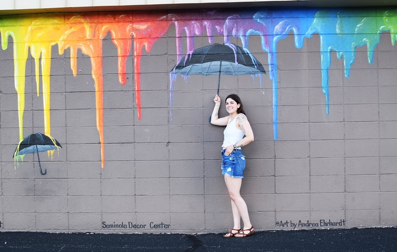 Tiffany stands in front of a gray wall with a mural of umbrellas and rainbow paint drips