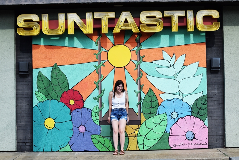 mural of flowers and a swing with a Suntastic sign above it, Tiffany 'sits' on the swing