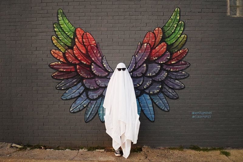 White sheet ghost stands in front of a gray wall with rainbow wings painted on it