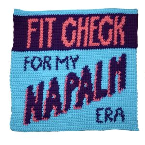 Crocheted tapestry piece in pink, light blue and dark purple reading ‘fit check for my napalm era’.